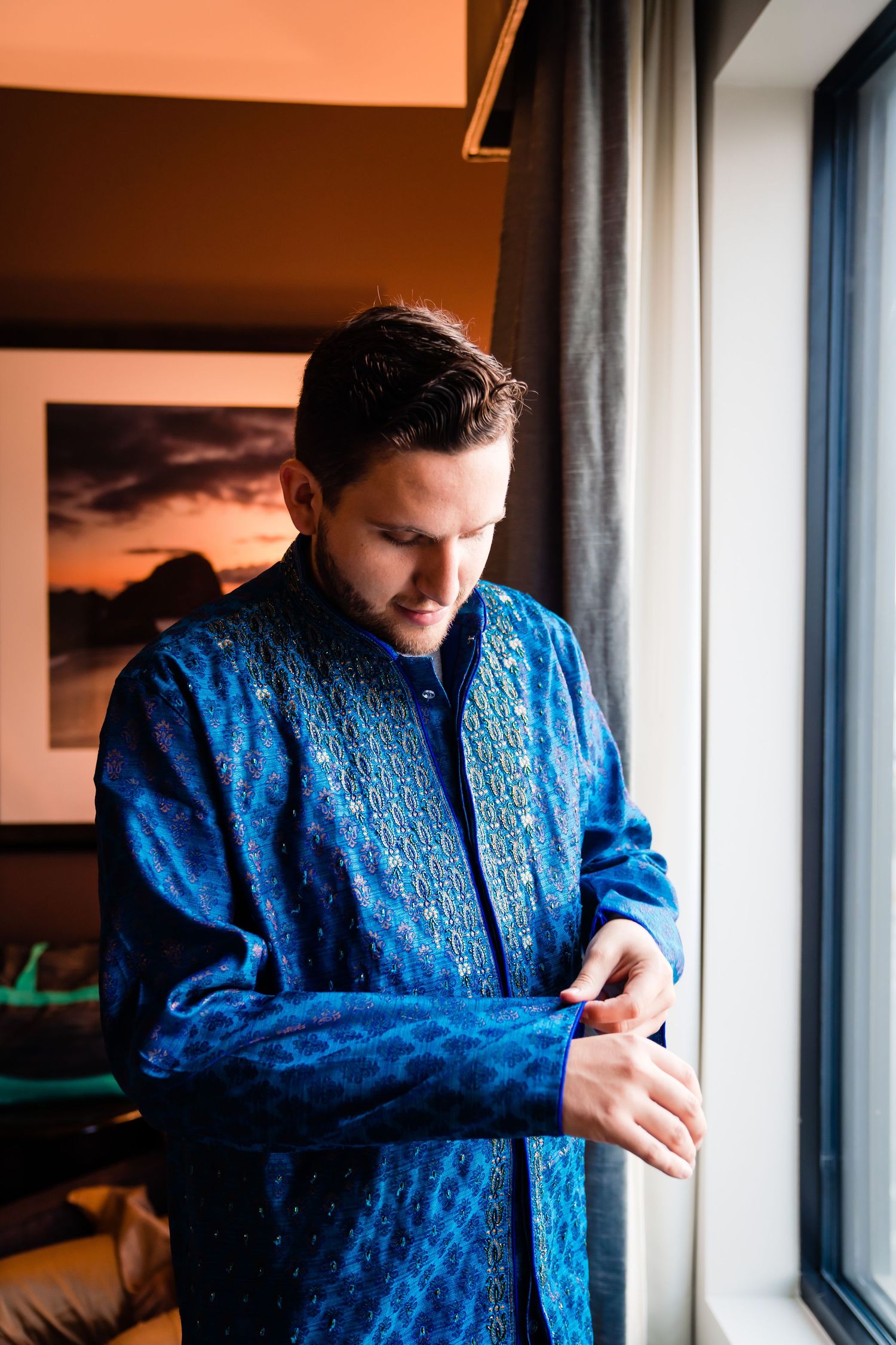 Person in blue patterned shirt looking down while standing near window in warm-lit interior space.