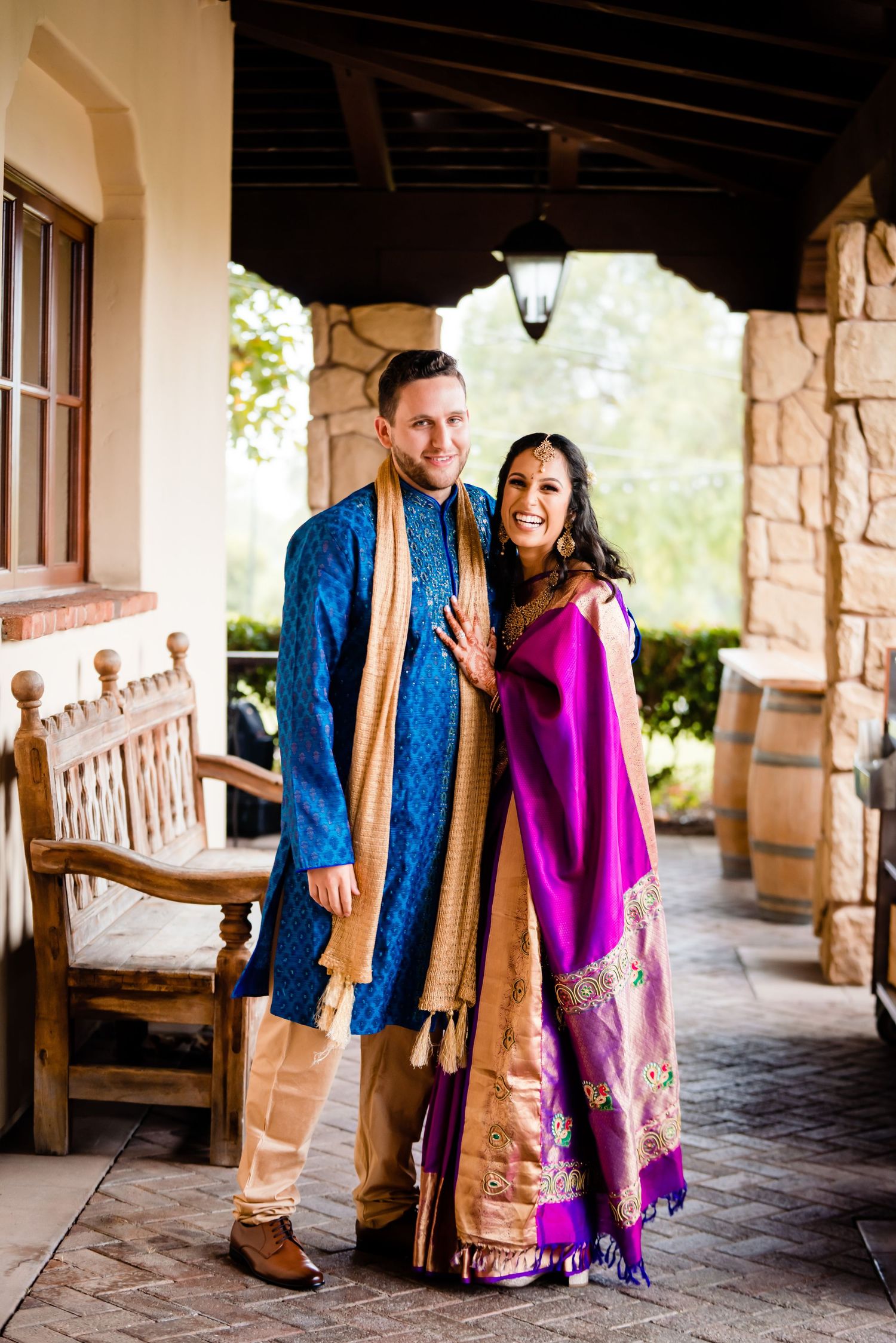 A couple in traditional Indian attire poses together under a stone archway with wooden benches in the background.