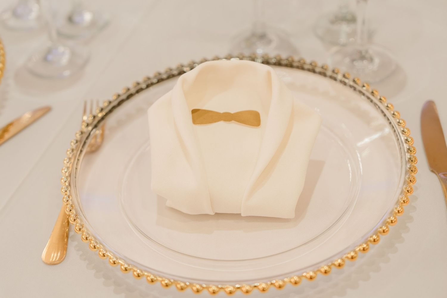 An elegant place setting with a white napkin and gold bow tie on a beaded charger plate.