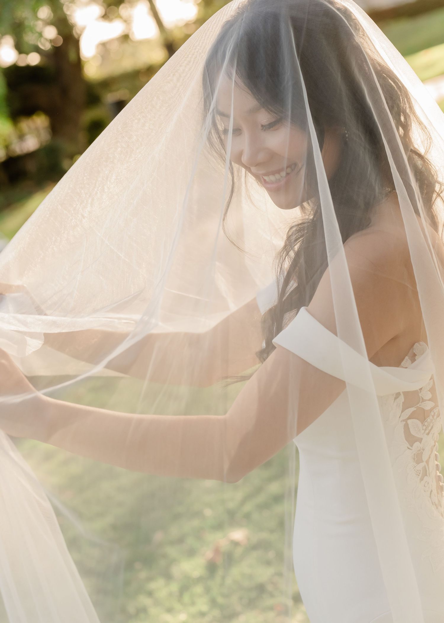 A bride in a flowing white veil poses gracefully outdoors in natural sunlight during a wedding photoshoot.