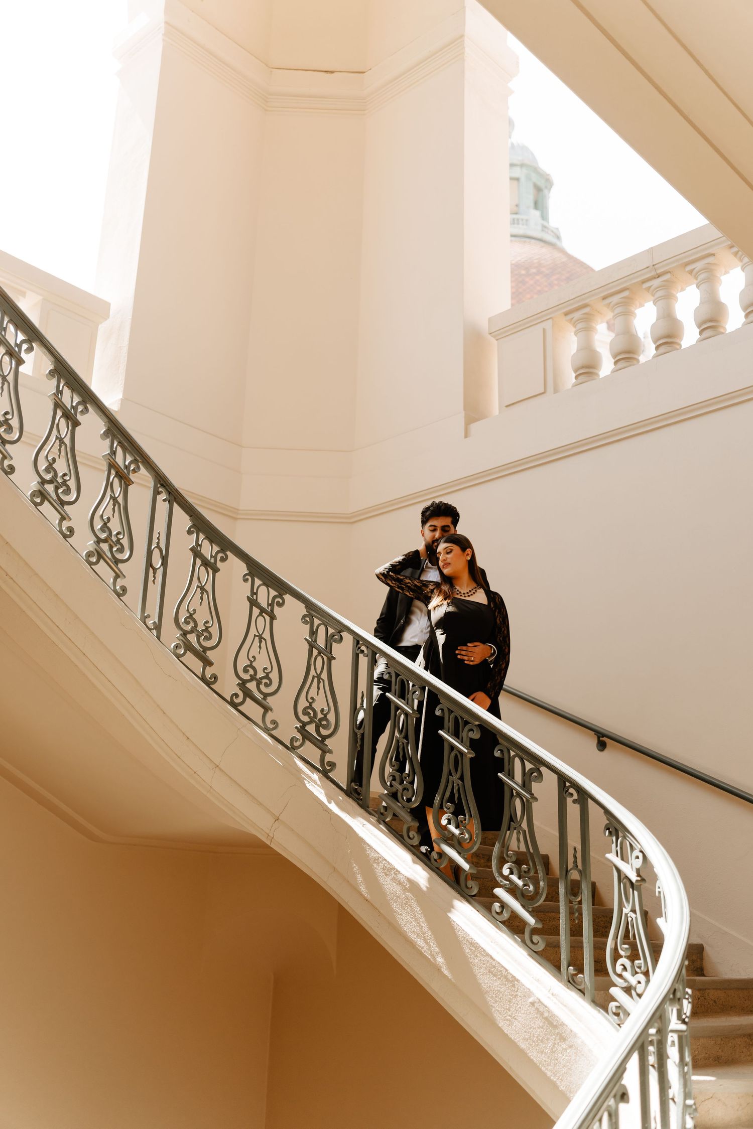 Couple embracing on elegant curved white staircase with ornate iron railing in a classical architecture building.