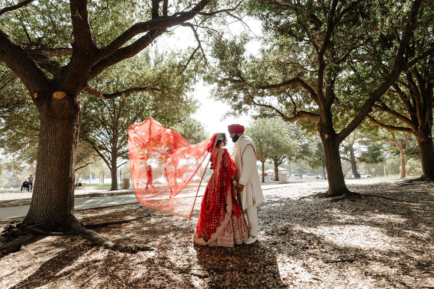 Indian bride in red lehenga and groom walk under oak trees, her dupatta flowing in the breeze during outdoor wedding.