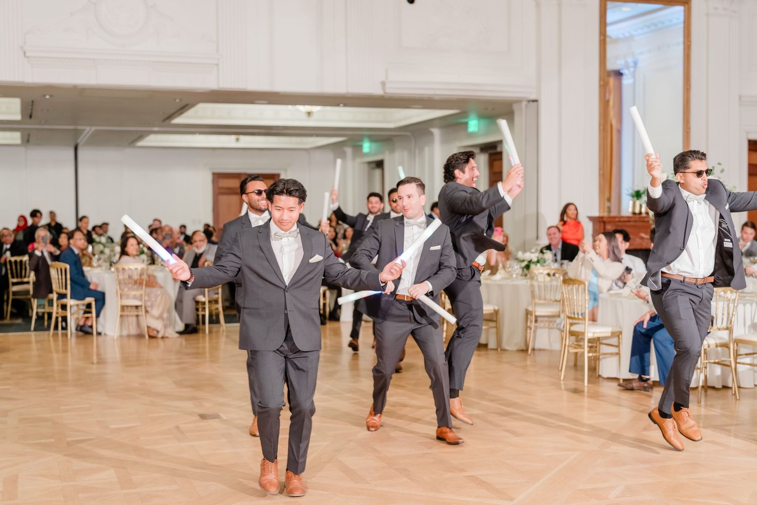 Group of groomsmen jumping and posing playfully for photos in a bright reception hall.