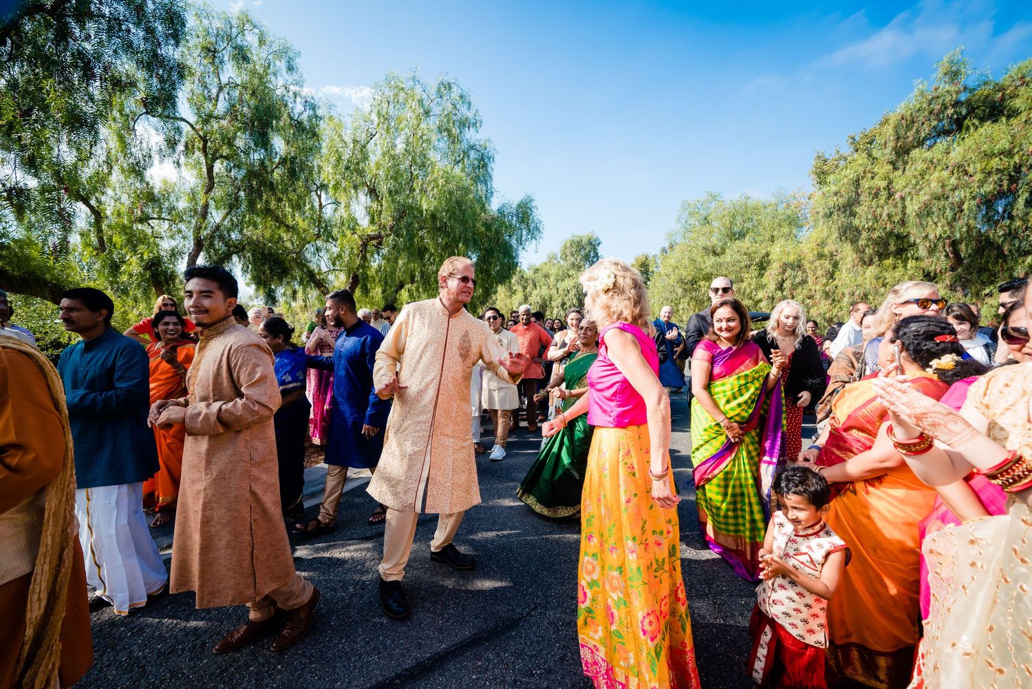 Colorful outdoor celebration with guests in traditional Indian attire walking together under trees on a sunny day.