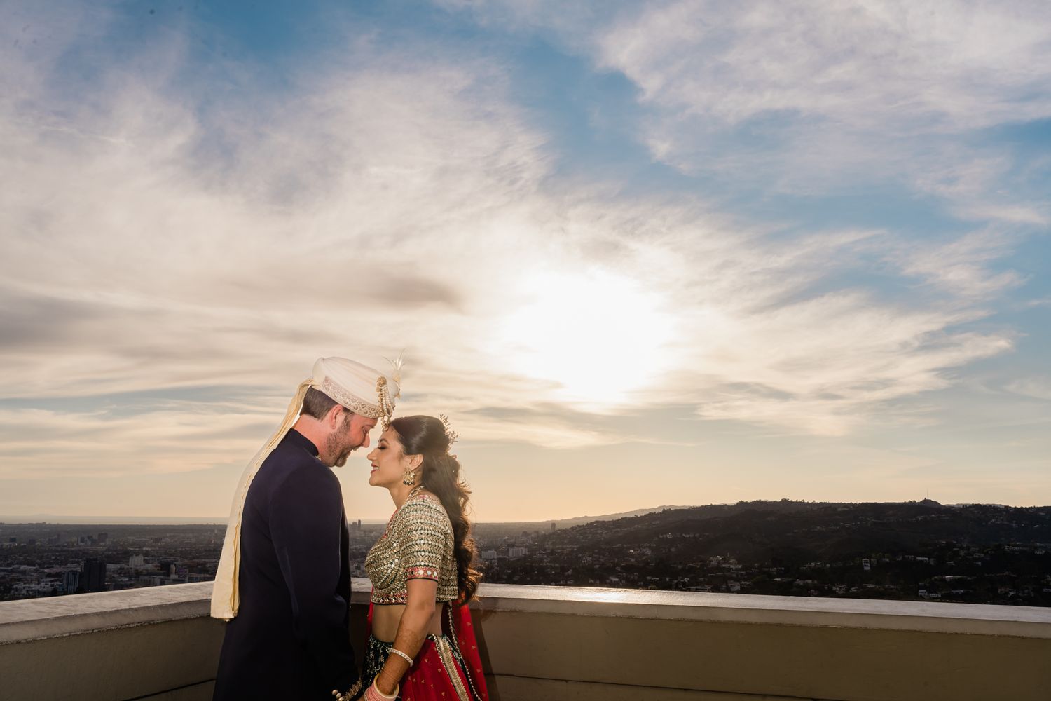 A couple shares a romantic moment at sunset on a rooftop with a scenic cityscape backdrop.