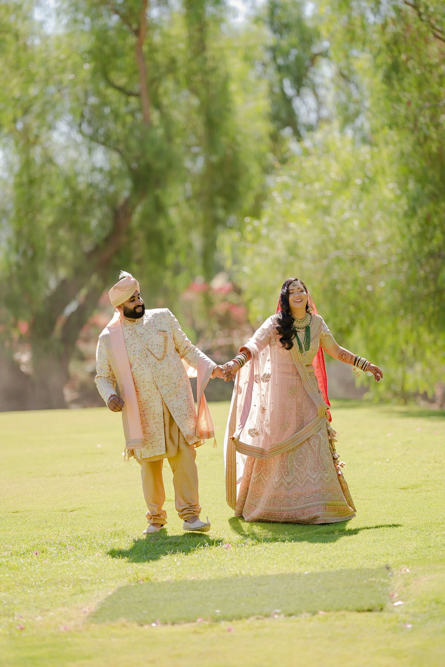 Couple in traditional Indian wedding attire walking together on a grassy lawn in a romantic outdoor setting.