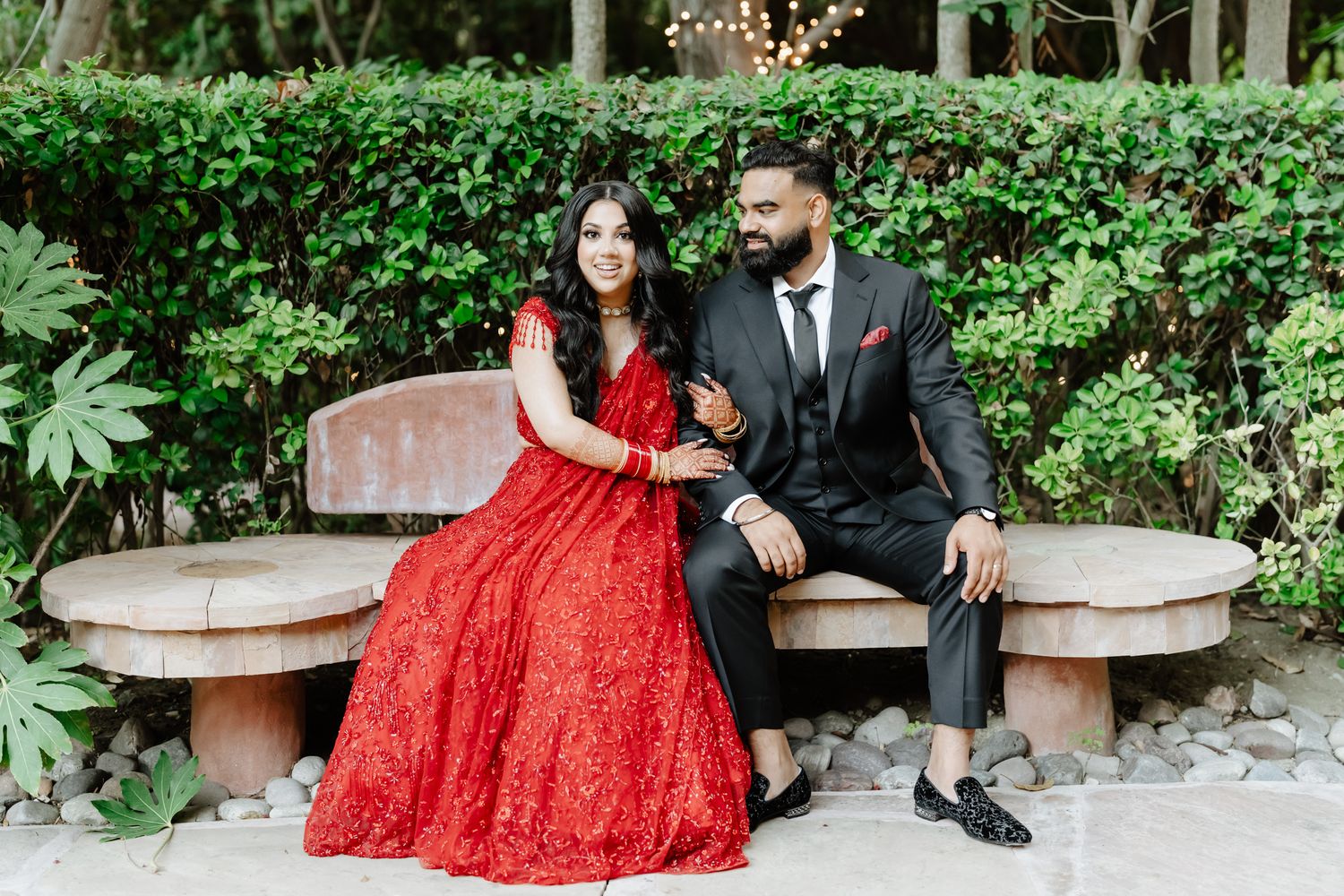 Couple in formal attire poses in a garden setting with the person wearing a stunning red lace gown.