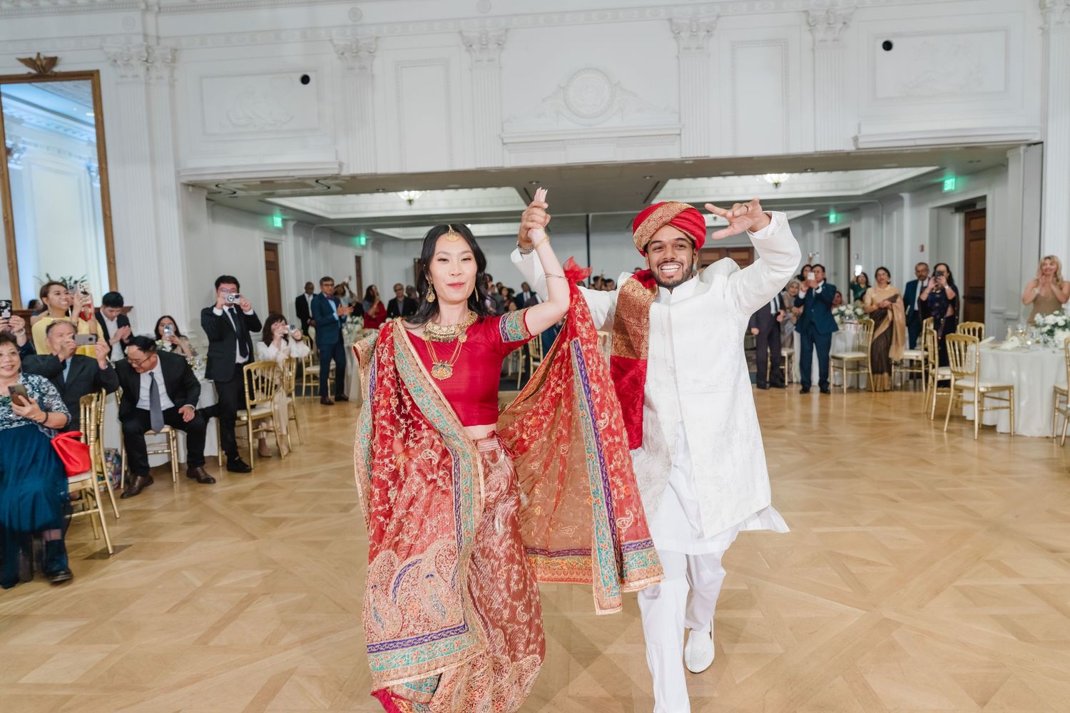 People in traditional Indian and Western formal wear dancing together in an ornate white ballroom.