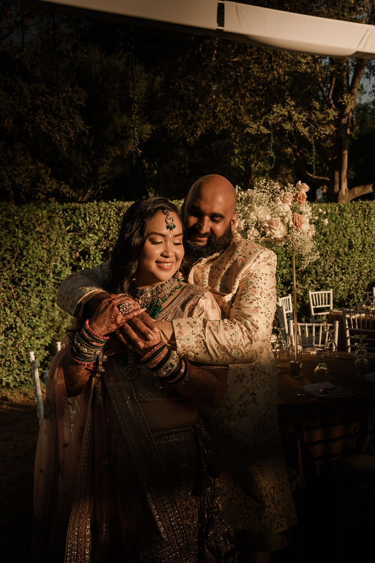 A couple in traditional Indian wedding attire embraces during golden hour in an outdoor garden setting.