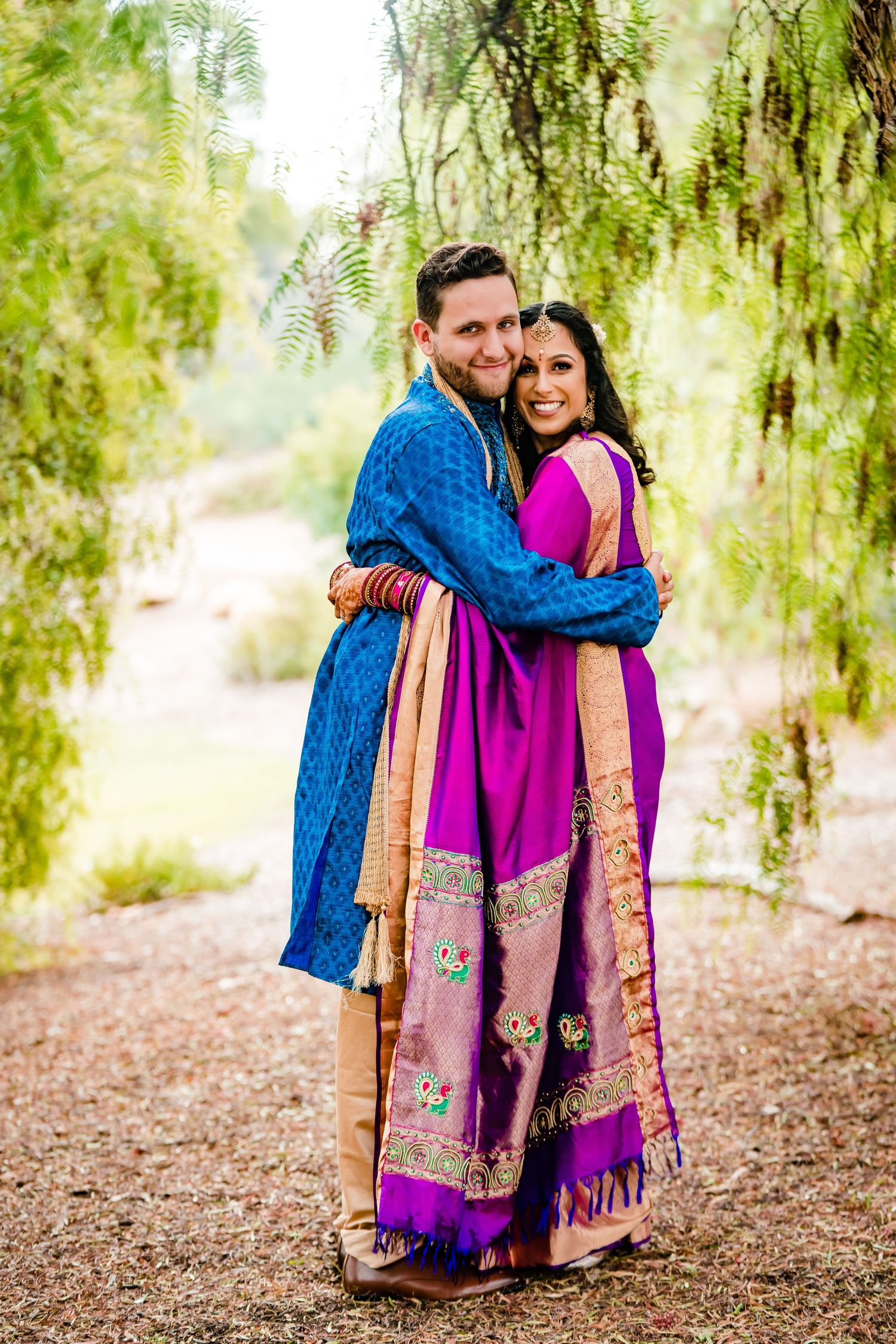 A couple in traditional Indian attire embrace under willow trees, with the woman wearing a bright magenta sari.