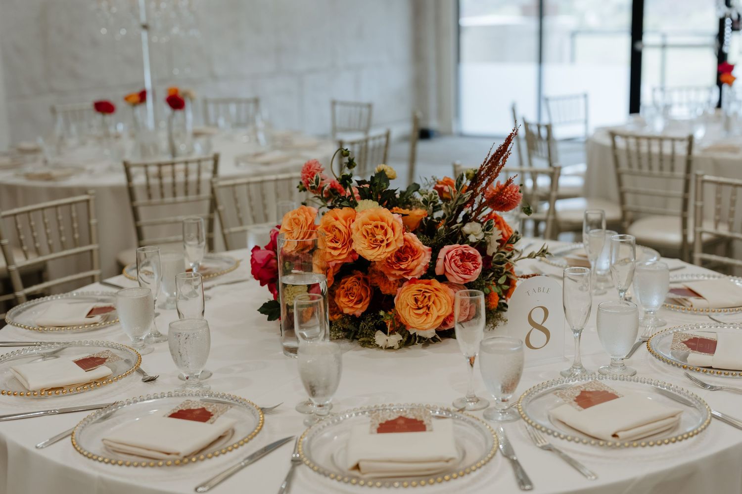 Elegant wedding reception table with orange and pink rose centerpiece, gold chiavari chairs, and white place settings.