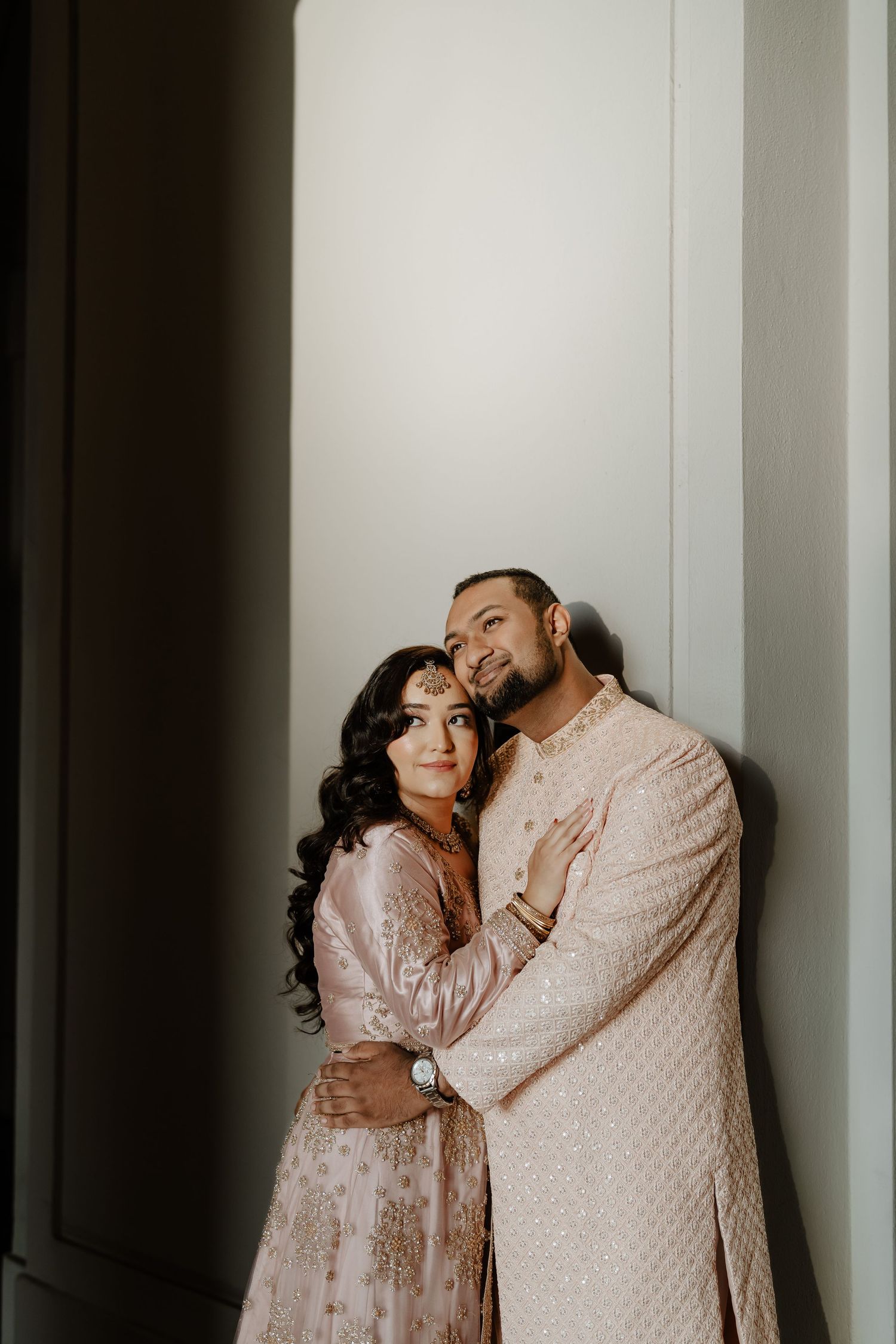 A couple in matching light pink and cream traditional Indian attire embrace lovingly against a white wall.