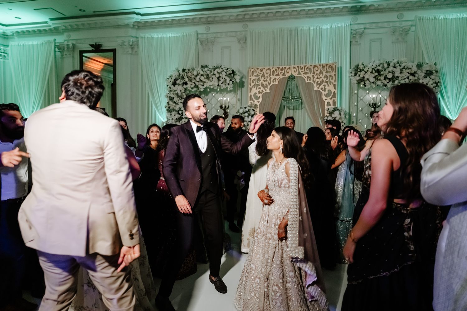 Wedding guests dance and celebrate on a glowing dance floor at an elegant reception venue with mint green lighting.