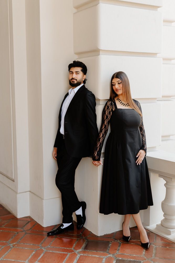 Elegant couple in matching black formal attire posing against a white architectural wall.