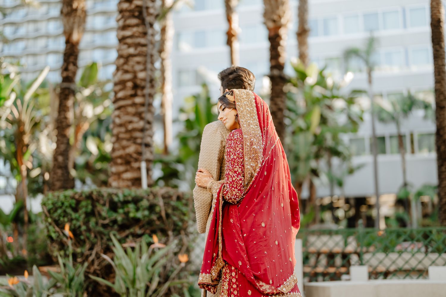 A person in a red and gold embroidered wedding sari poses among tropical palm trees.