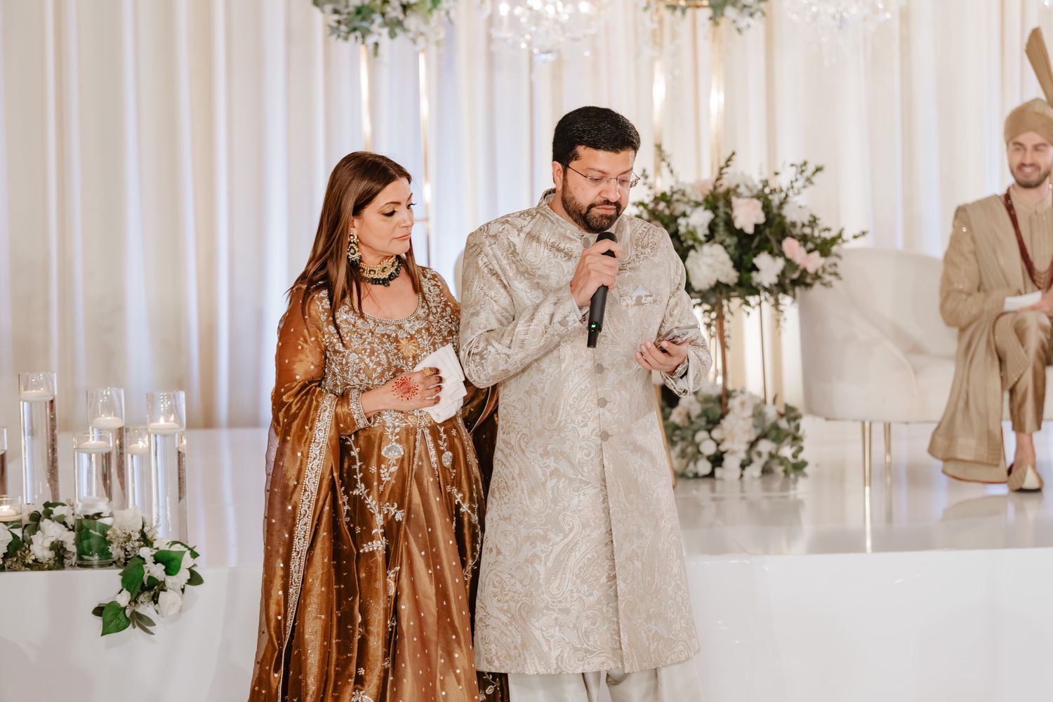 A couple in traditional South Asian wedding attire speaks at a reception decorated with white floral arrangements.