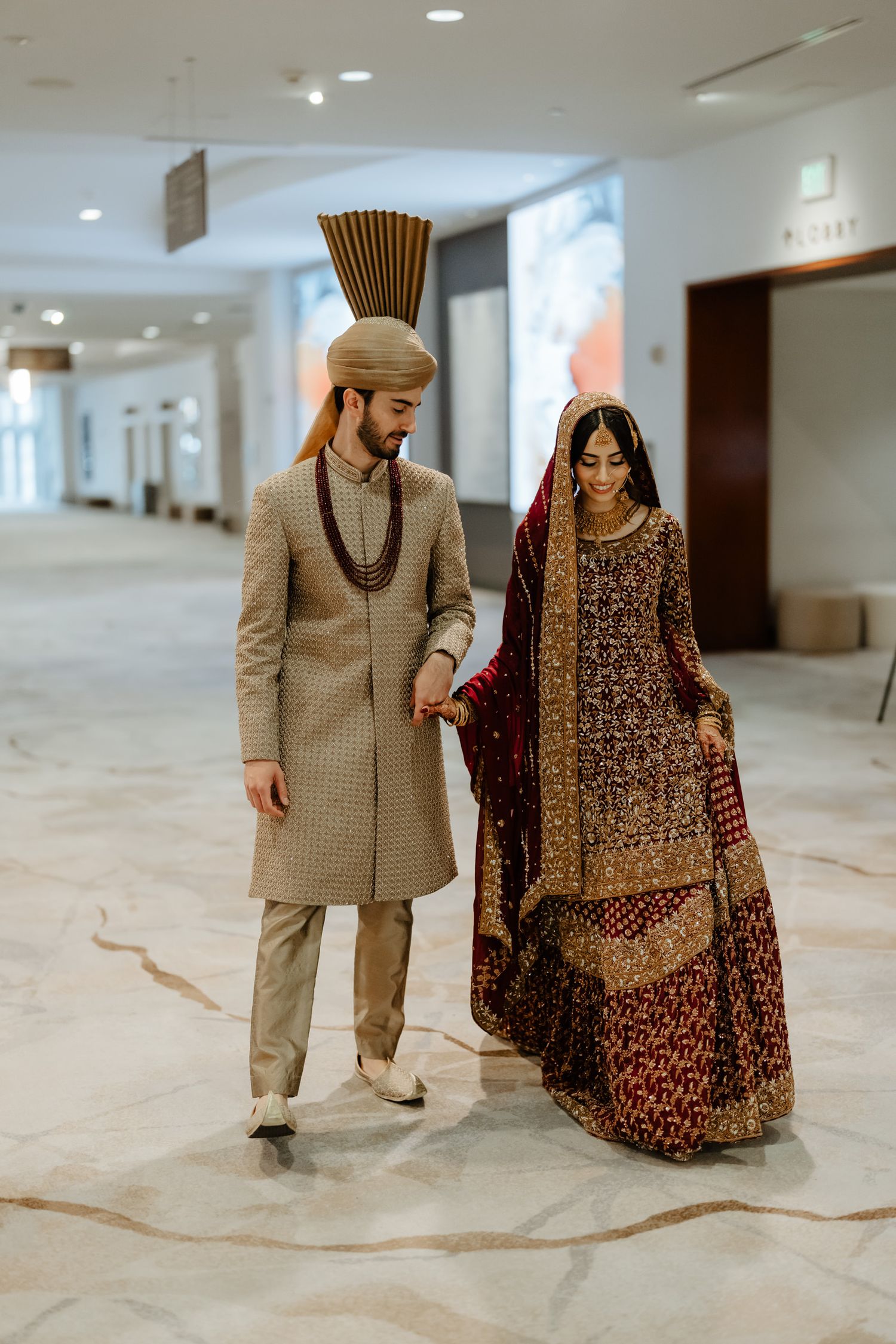 A couple in traditional South Asian wedding attire walking together in a white marble hallway.
