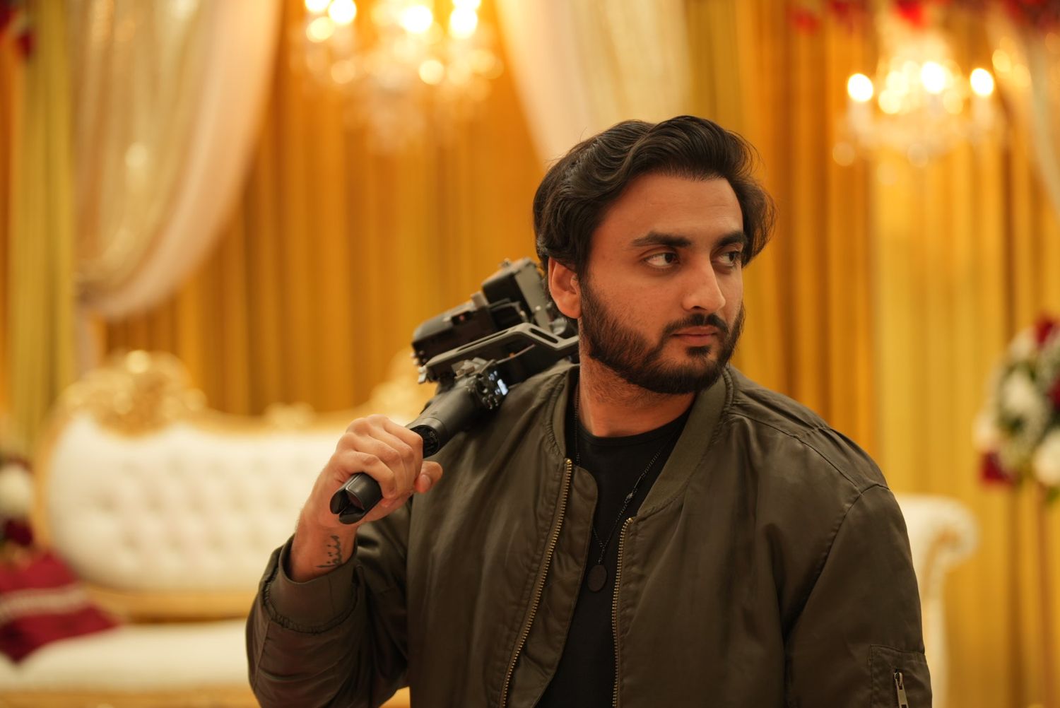 Man in olive jacket holds a camera at a decorated indoor venue with golden drapes.