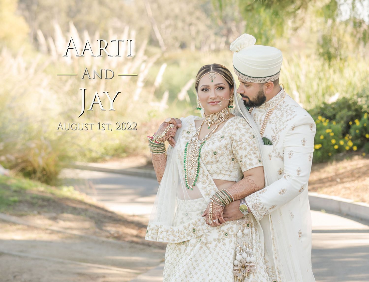 A couple in traditional white and gold Indian wedding attire poses together in a sunlit outdoor garden setting.