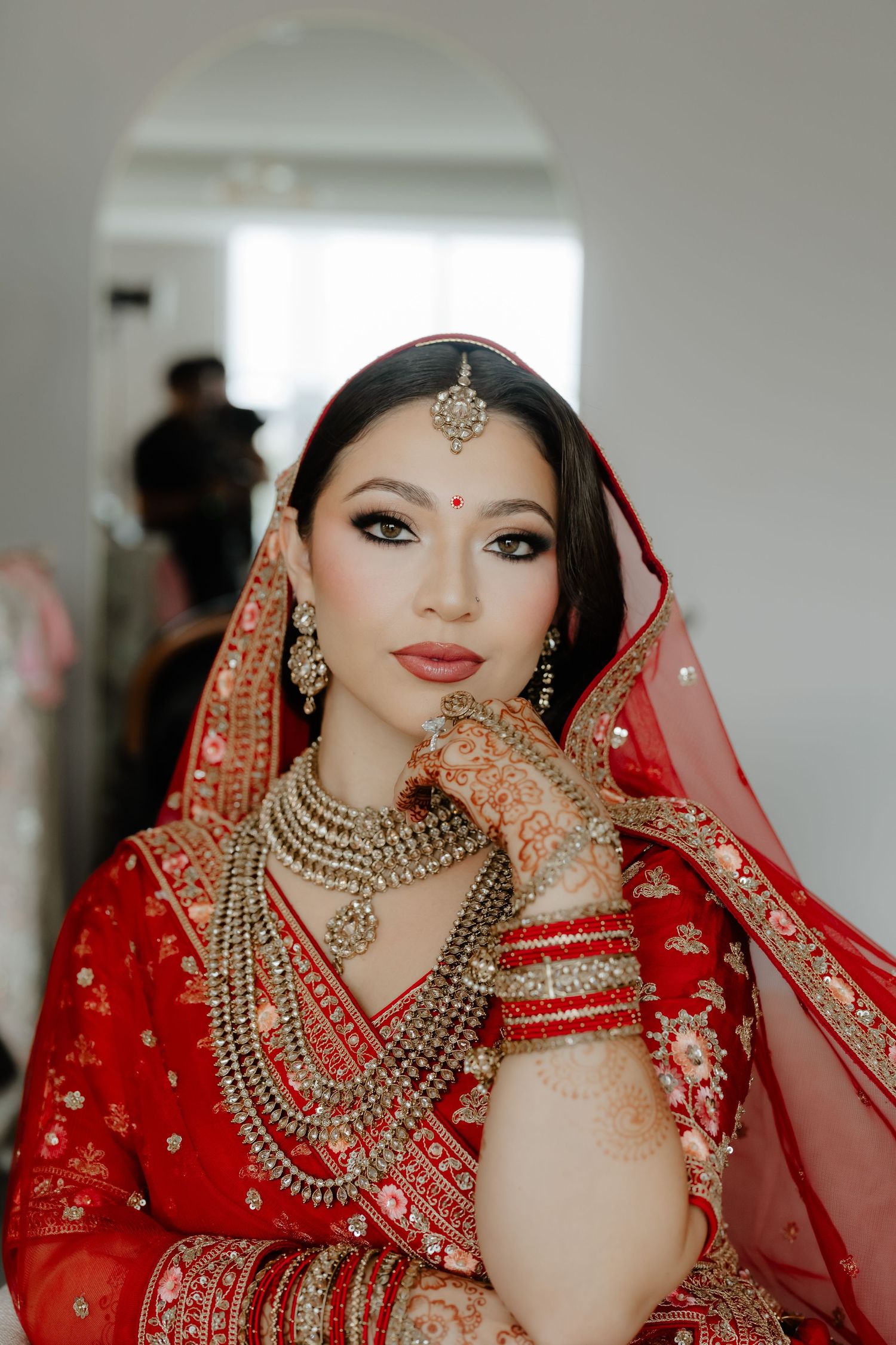 Indian bride in red lehenga with gold jewelry, maang tikka, bangles, and henna hands poses elegantly for bridal portrait.