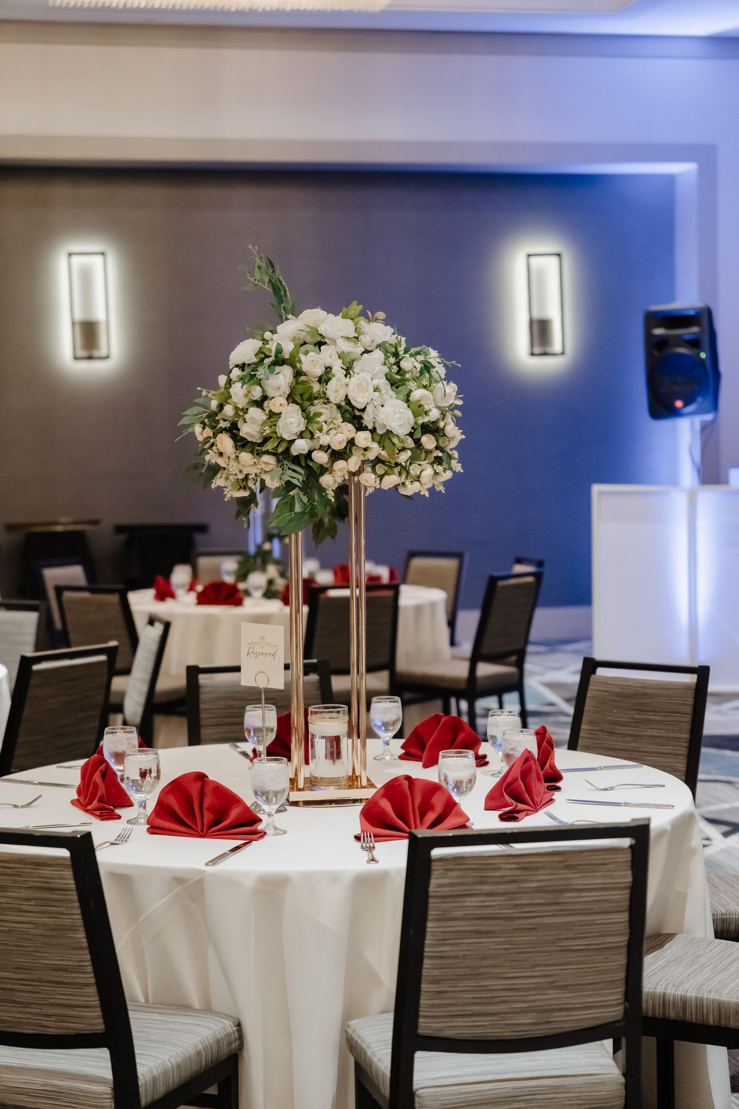 Elegant wedding reception table with tall white floral centerpiece and red napkins folded in a formal dining setting.