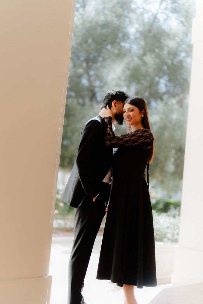 Romantic couple in matching black formal attire embracing between white columns outdoors.
