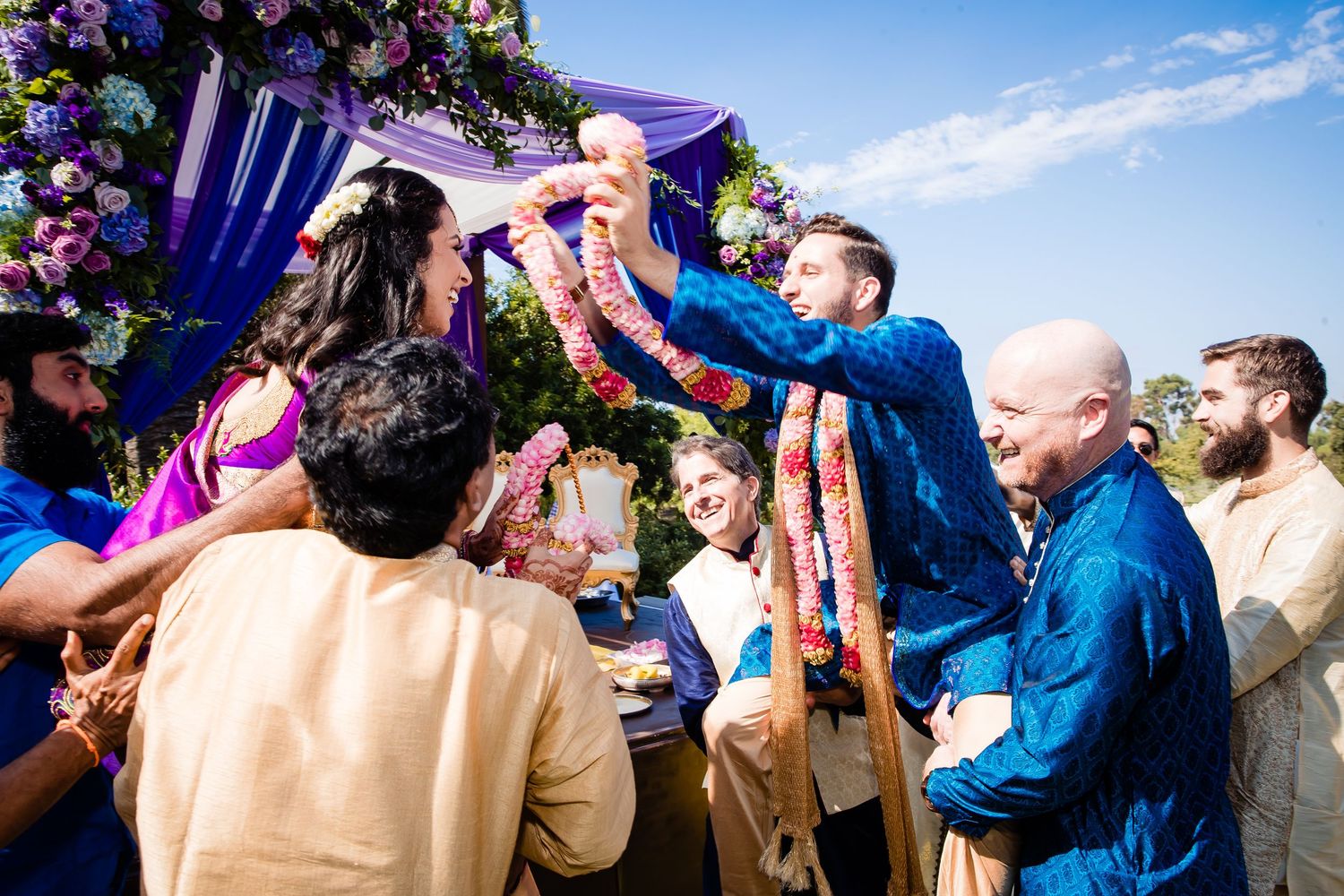 Wedding guests dance and celebrate together under a blue sky at an outdoor reception.