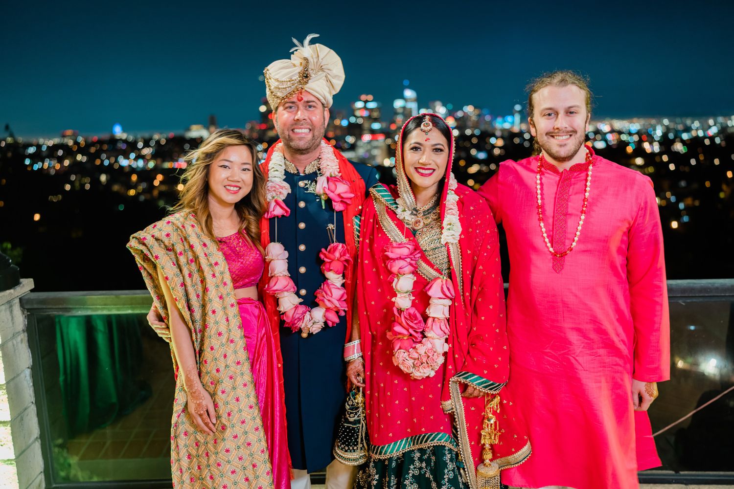 A group in festive attire poses together at a nighttime outdoor event with city lights in the background.