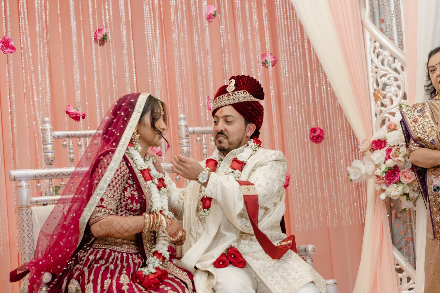 A couple in traditional Indian wedding attire performs a ceremony ritual as rose petals fall around them.