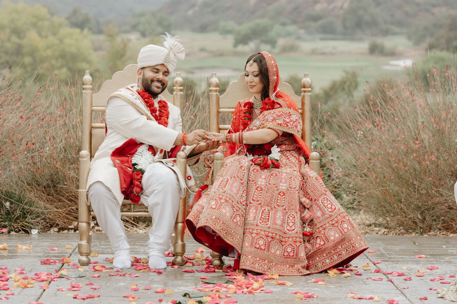 Indian bride and groom in traditional red and white wedding attire seated on golden chairs during outdoor ceremony.