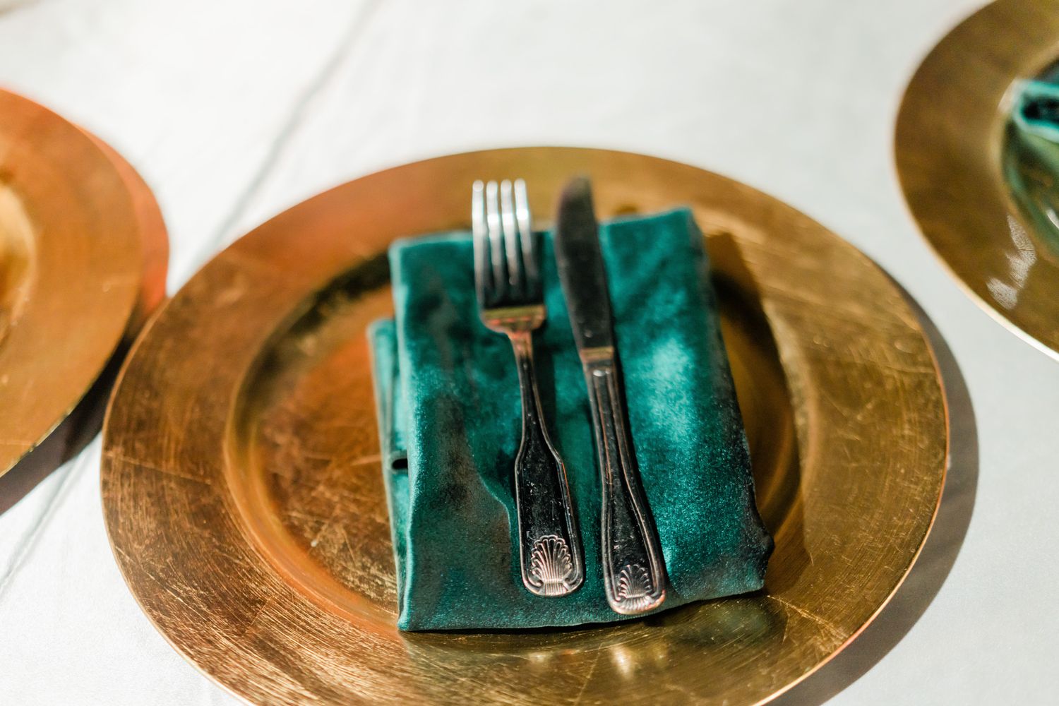 Gold charger plates with teal napkins and silverware arranged in sequence on a white surface.