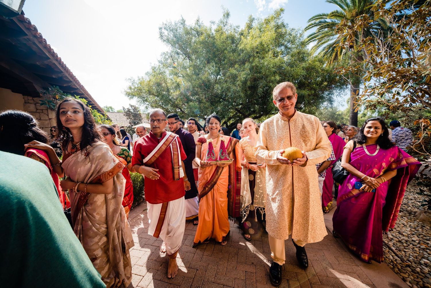 Wedding guests dance and celebrate together outdoors with vibrant orange and pink attire at a joyful reception.