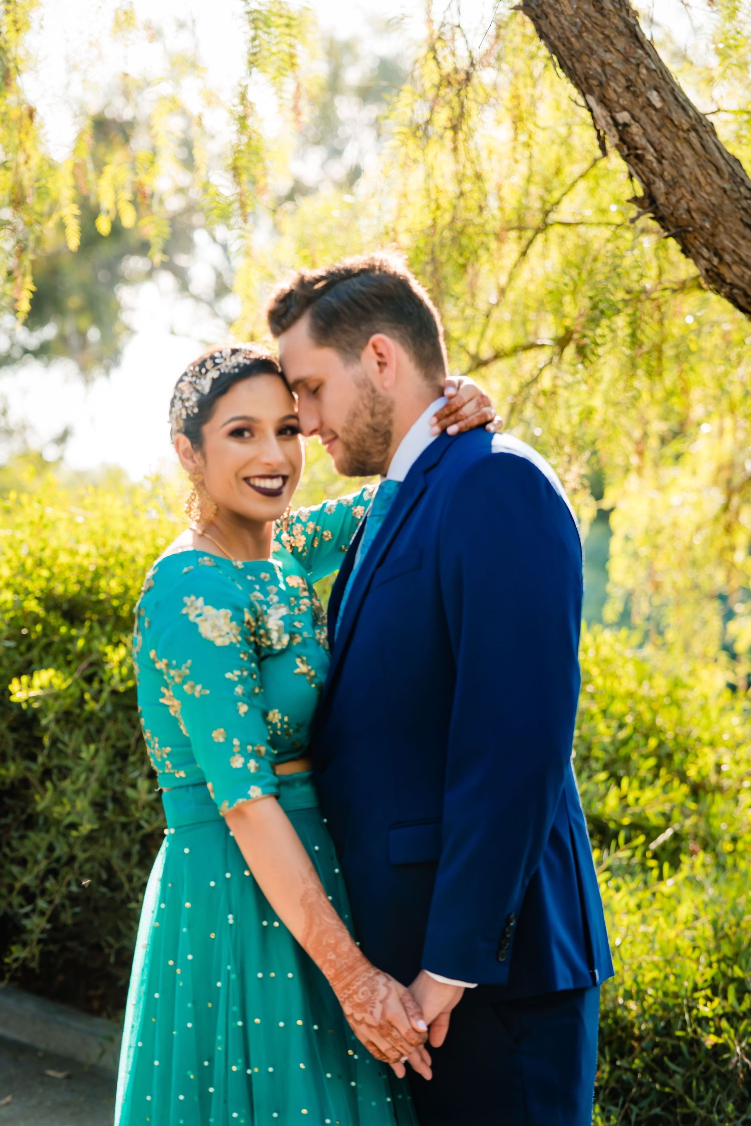 Couple in blue navy suit and teal dress share romantic moments under willow trees in golden sunlight.