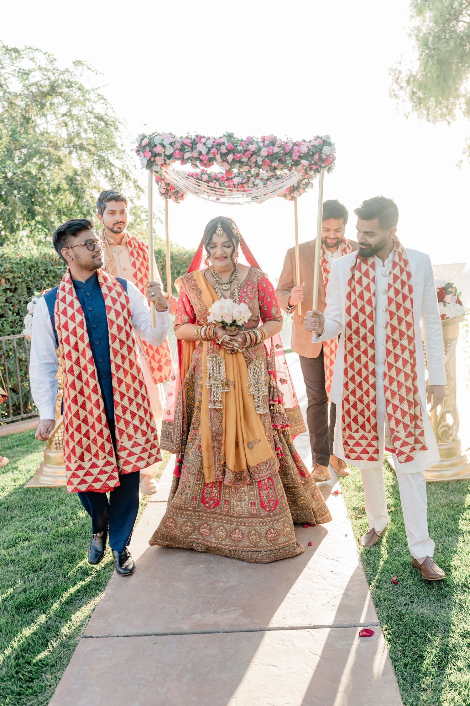 A traditional Indian wedding procession with attendees wearing red and white patterned shawls walking under a floral canopy.