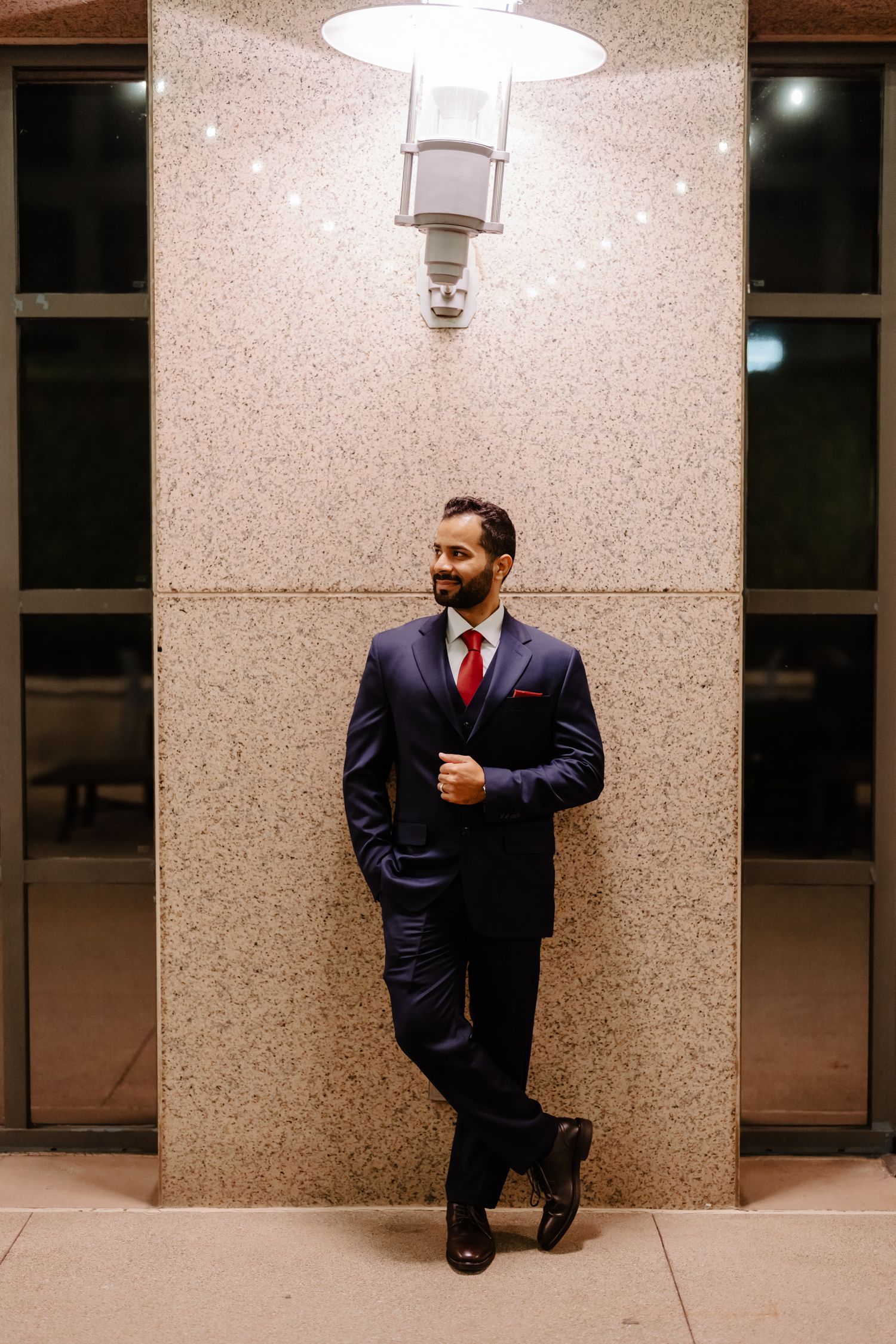 Formal portrait under lighting featuring navy suit and red tie against textured wall.