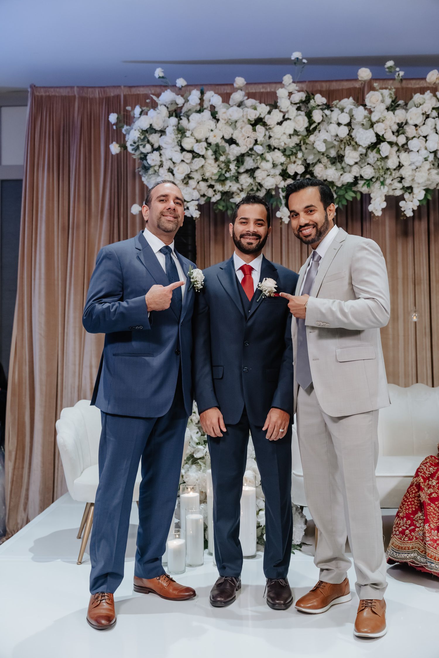Three men in suits pose together at a wedding ceremony under an elegant white floral arch display.