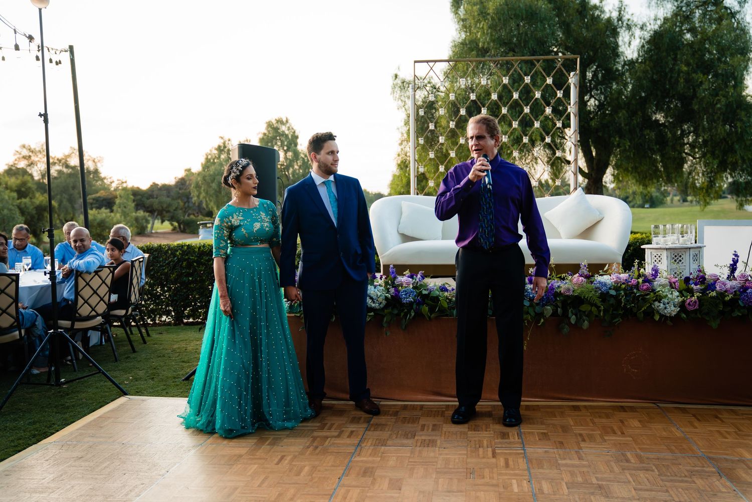 People in formal attire gather at an outdoor wedding reception on a patio decorated with string lights at sunset.
