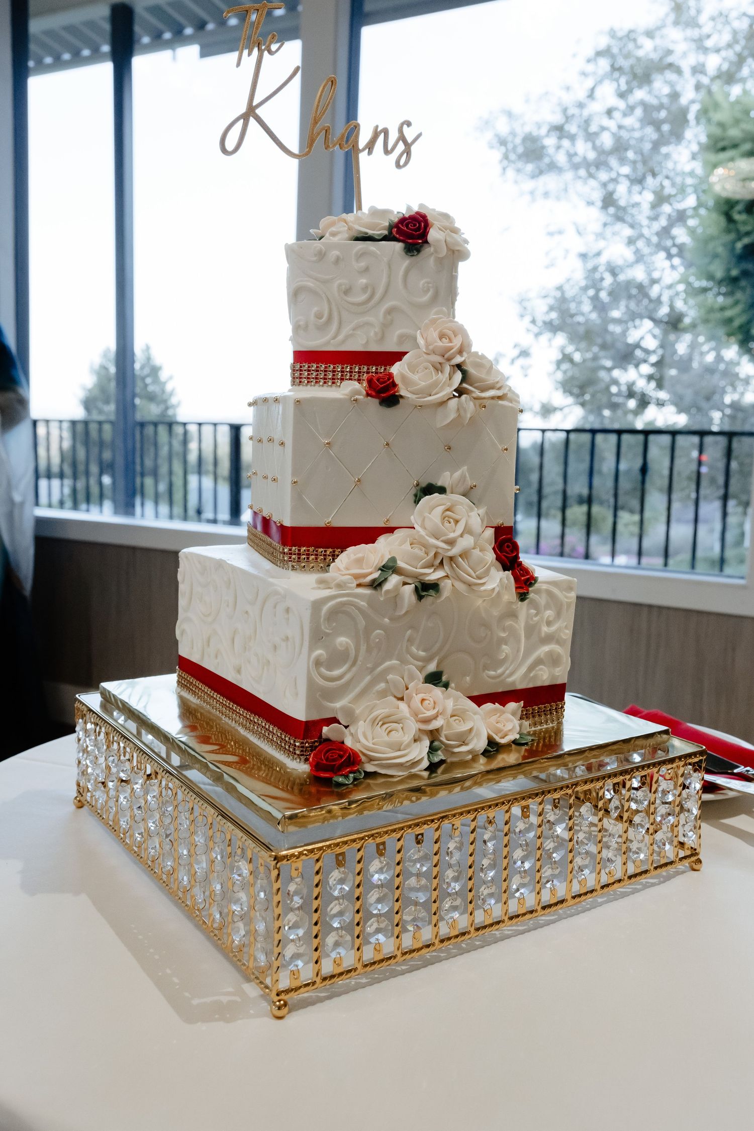 Elegant four-tier white wedding cake with red roses, gold trim, and crystal stand, topped with The Khans cake topper.