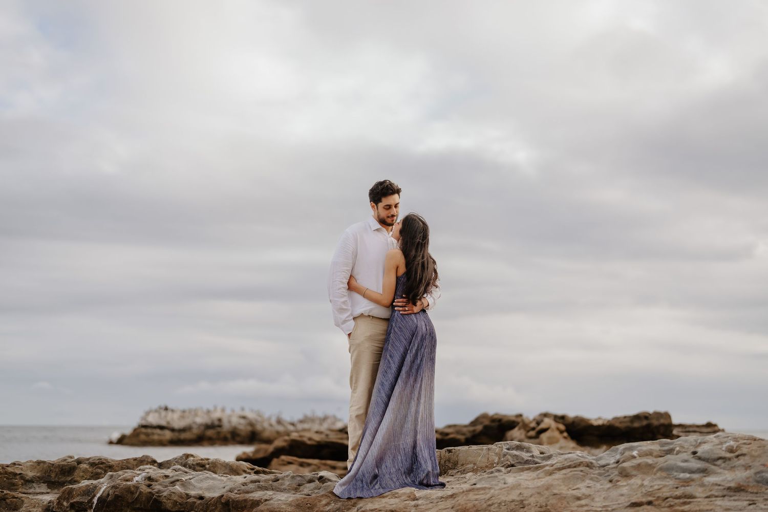 Couple standing on rocky coastal shore, woman in flowing purple dress, man in white shirt, overcast sky backdrop.