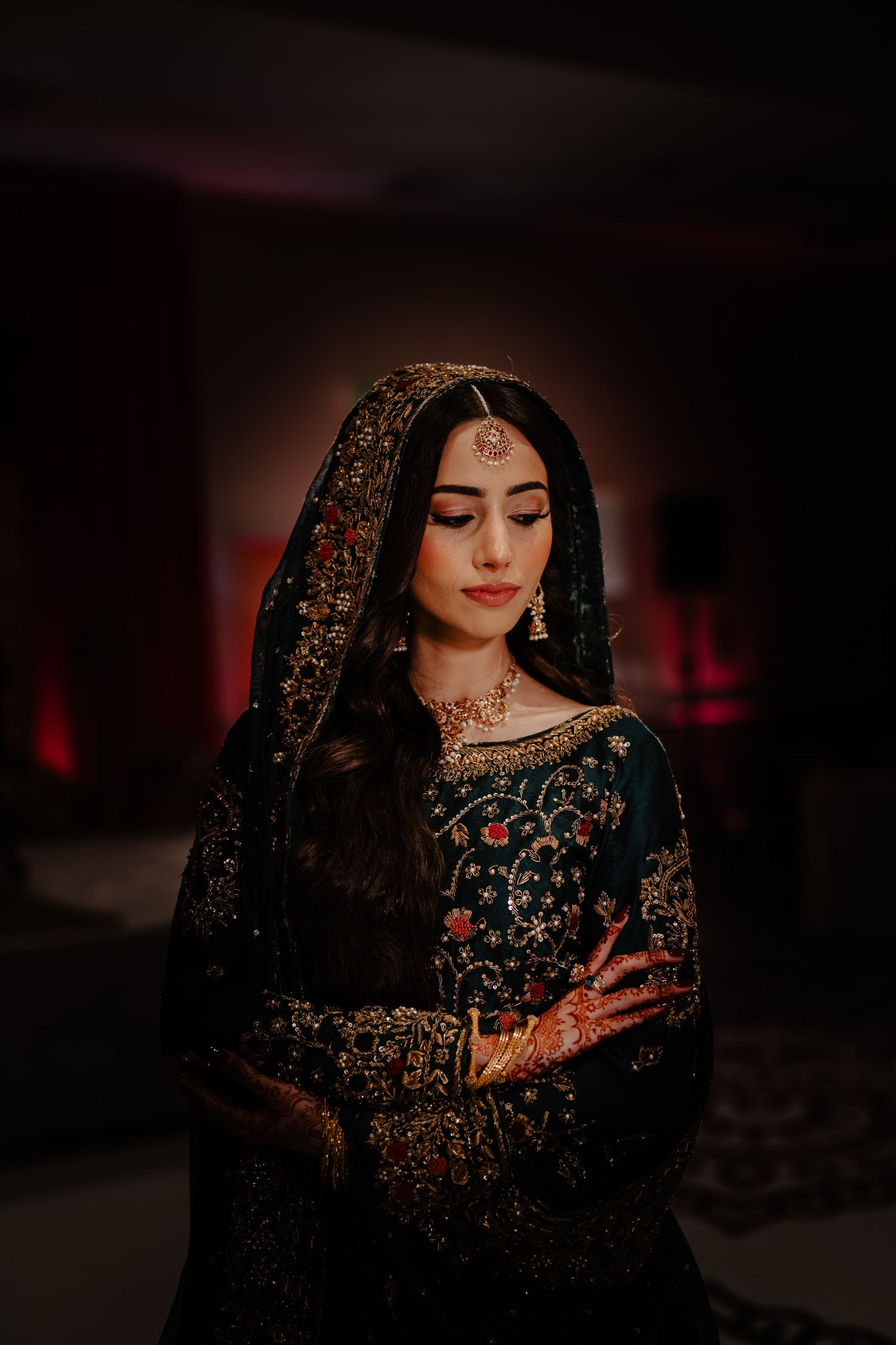 Traditional South Asian bride wearing an ornate black and gold embroidered dress with henna adorned hands in moody lighting.