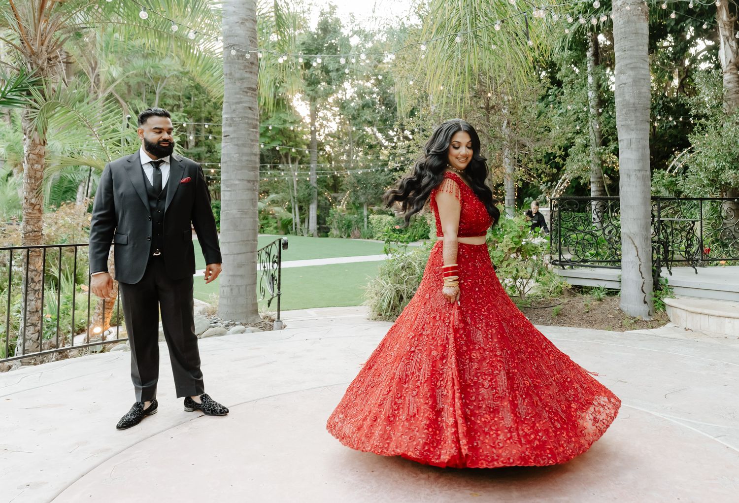 A couple in formal attire pose separately outdoors, with the bride twirling in a sparkly red lehenga.