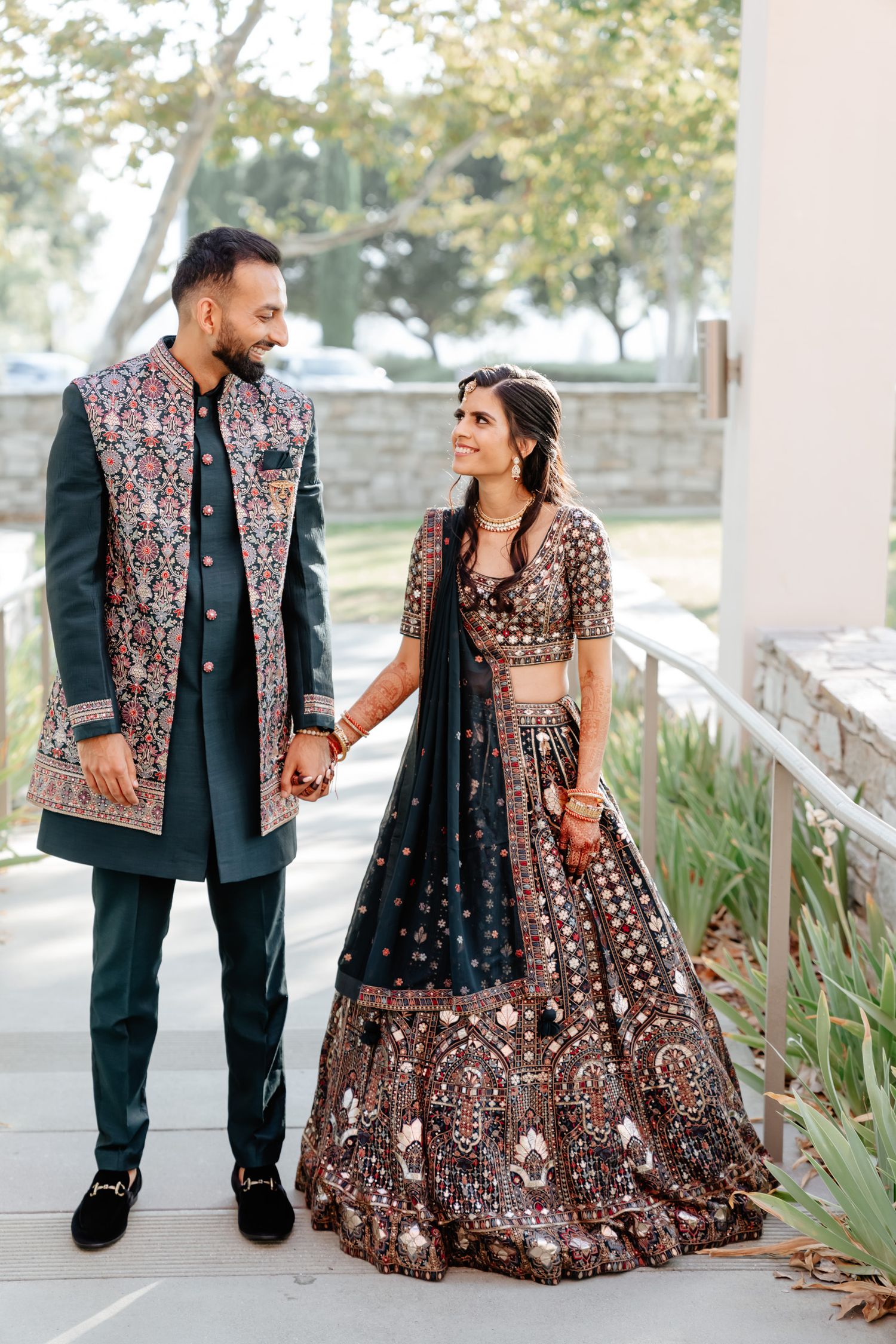 Couple in coordinated Indian wedding outfits walking along a garden pathway.