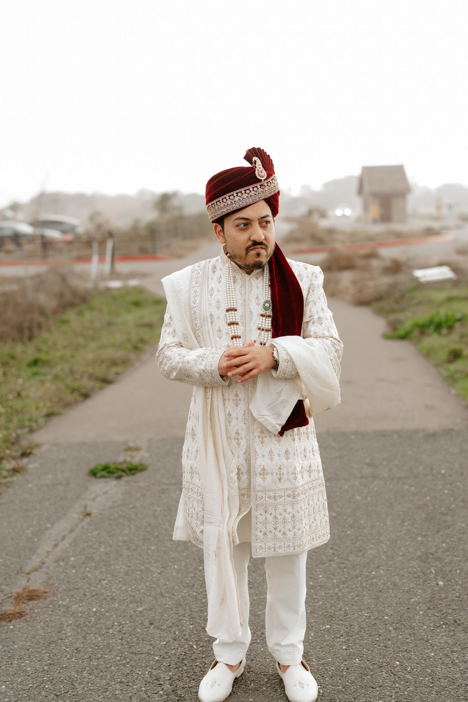 Traditional South Asian groom wearing white embroidered sherwani and maroon turban poses outdoors on a path.
