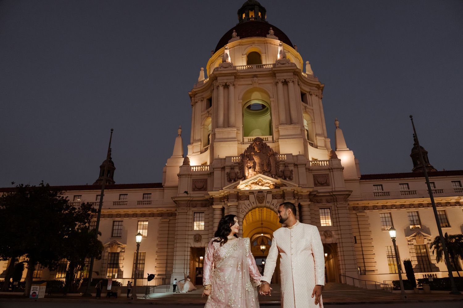 A couple in traditional wedding attire pose in front of an illuminated Pasadena City Hall at night.