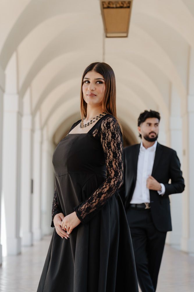 Woman in black lace-sleeve gown poses in elegant arched hallway with suited man in background.