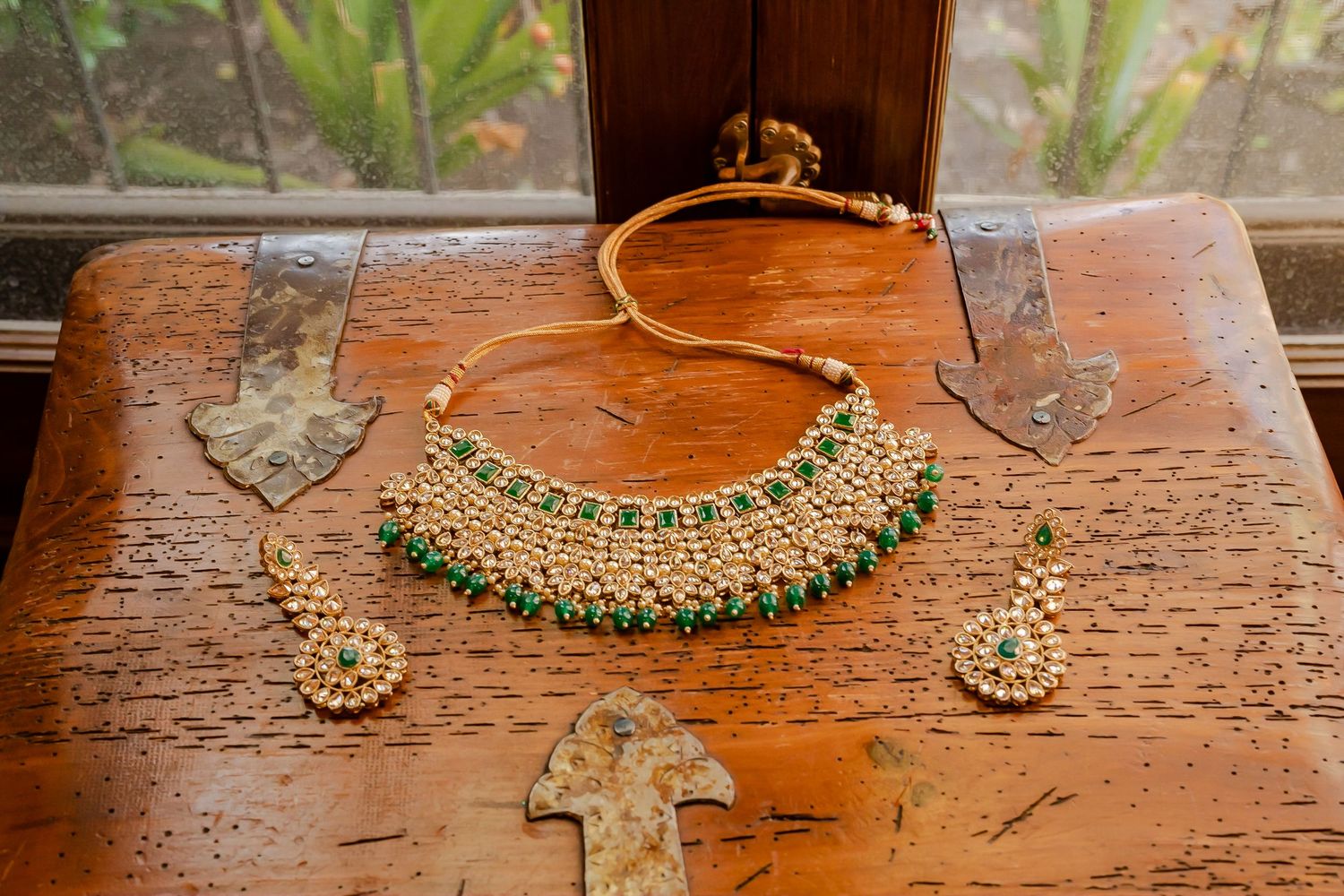 Ornate gold and emerald Indian jewelry pieces including necklaces and earrings displayed on a rustic wooden surface.