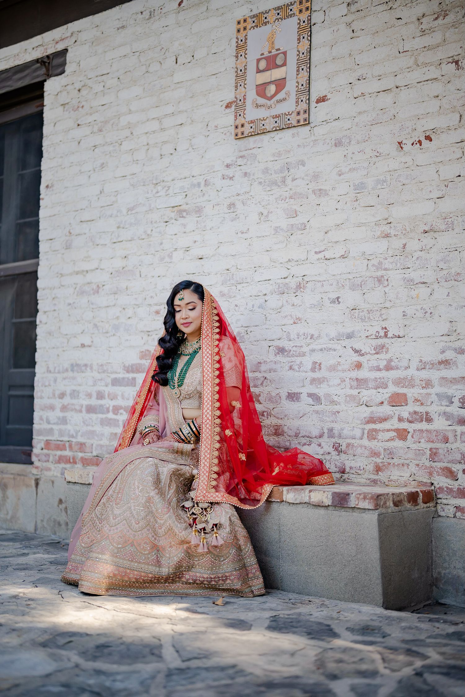Traditional Indian bride in red and gold lehenga sitting against white brick wall.