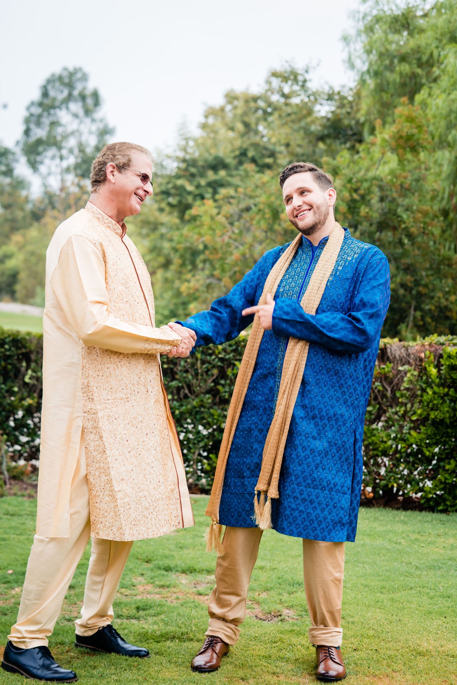 Two men in traditional South Asian attire share a playful moment on a green lawn, one in peach and one in blue.