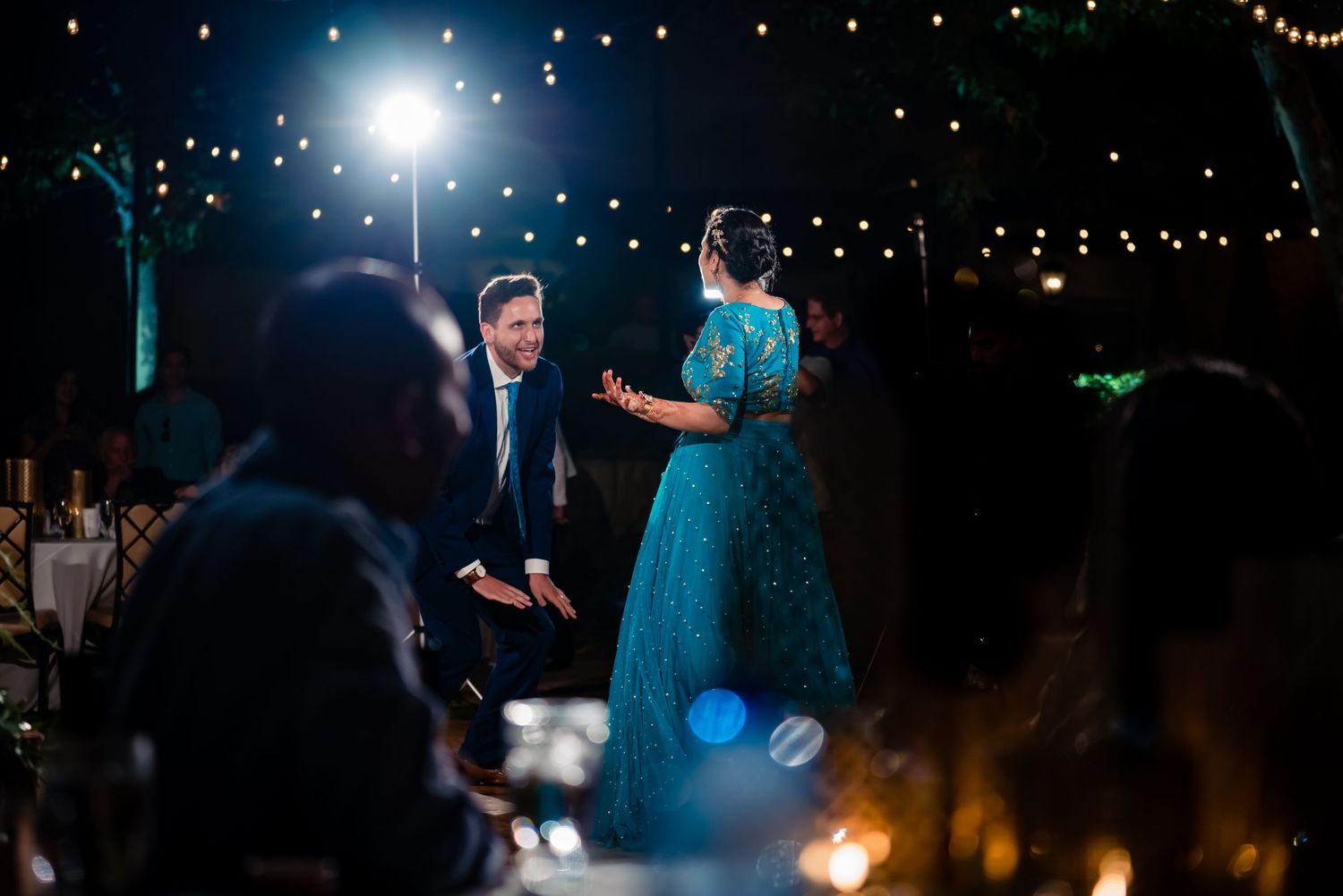 A couple dances together at a dimly lit outdoor wedding reception with string lights overhead.