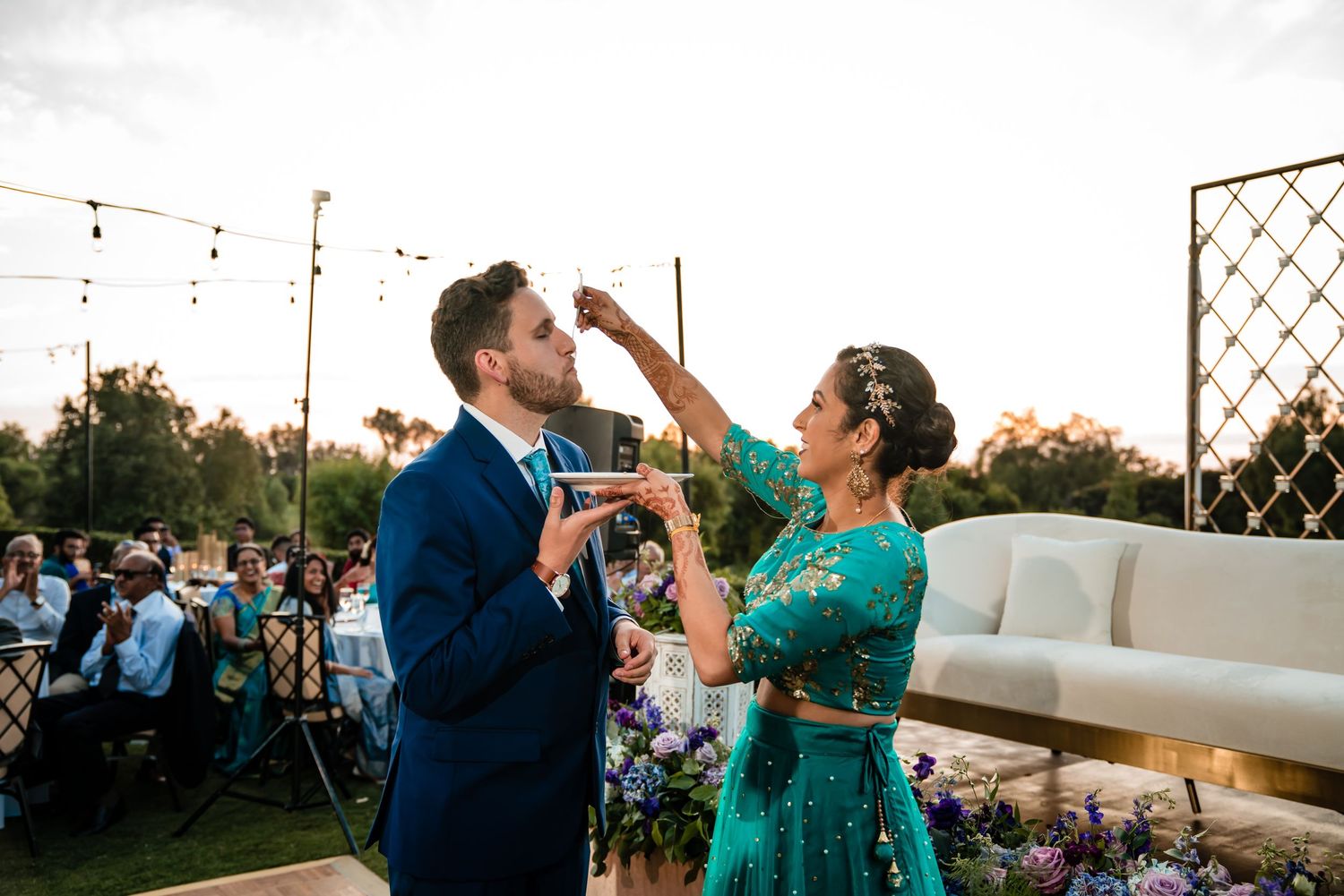 A couple in formal attire performs a celebratory toast at an outdoor wedding reception during sunset.